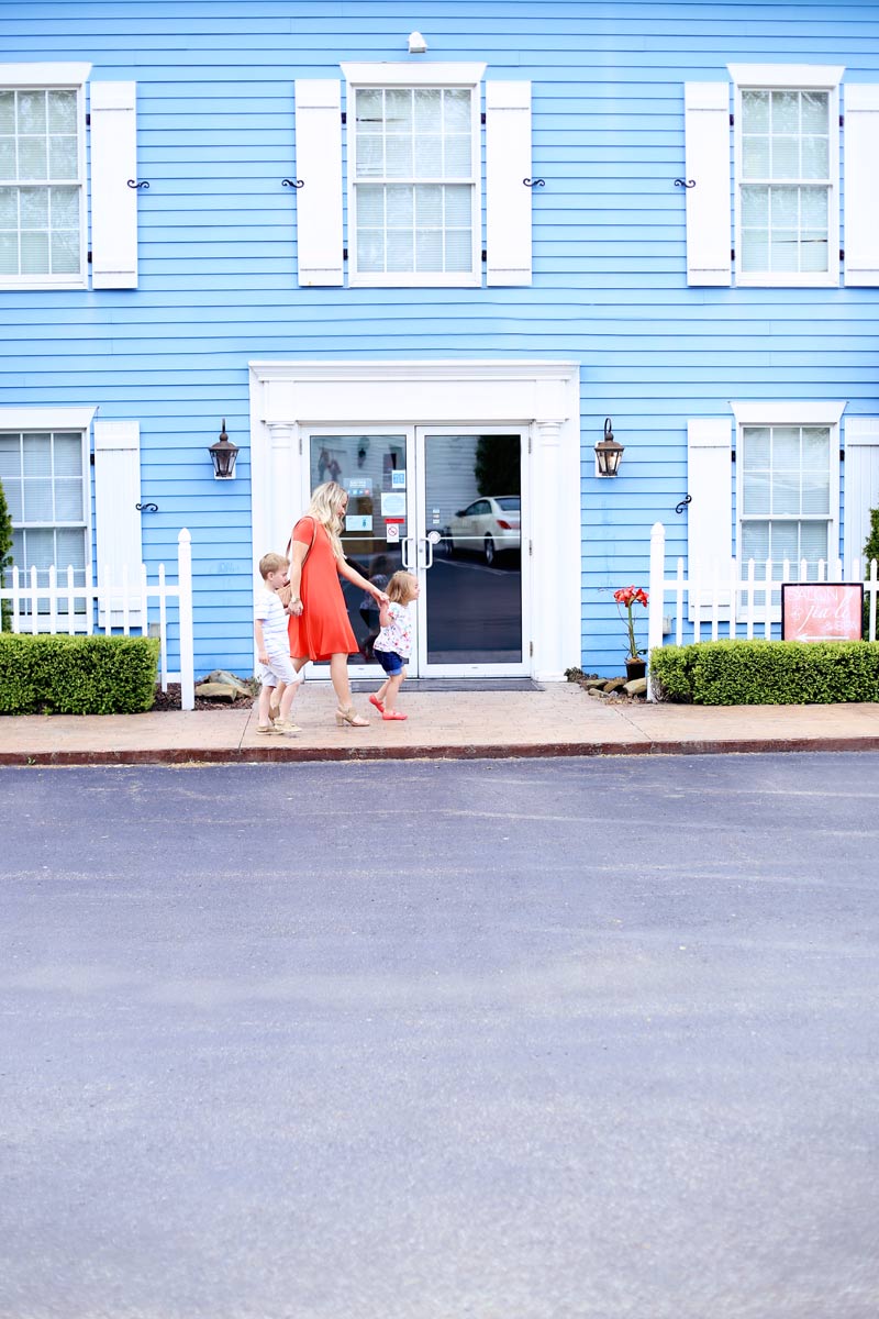Abby wears a red dress and walks hand in hand with her children as they pass the entrance of a bright blue house.